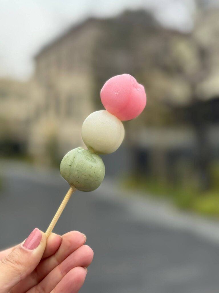 Fotografia autoral de um espetinho de feijão doce japonês (Dango) colorido, com bolinhas nas cores rosa, branco e verde, servido em um espetinho de madeira minimalista, ilustrando a estética do respeito e a simplicidade na culinária do Japão.