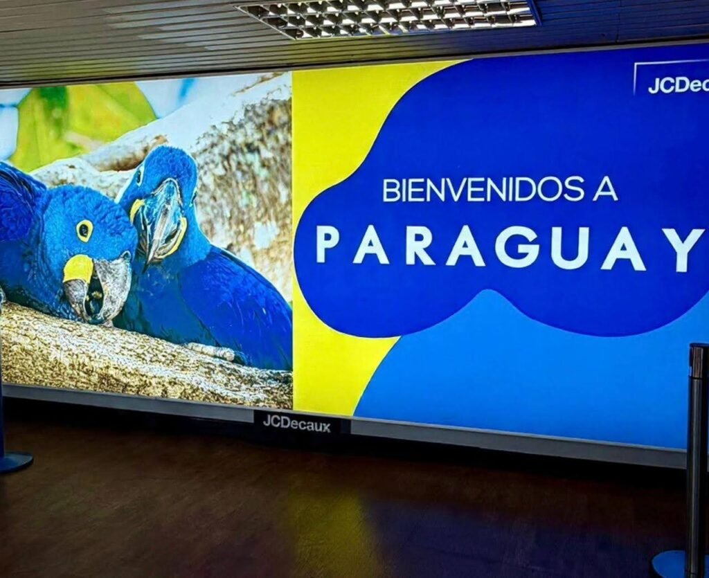 Mesa de recepção em evento de negócios no Paraguai, com telão ao fundo exibindo duas araras azuis e o texto Bienvenidos a Paraguai.