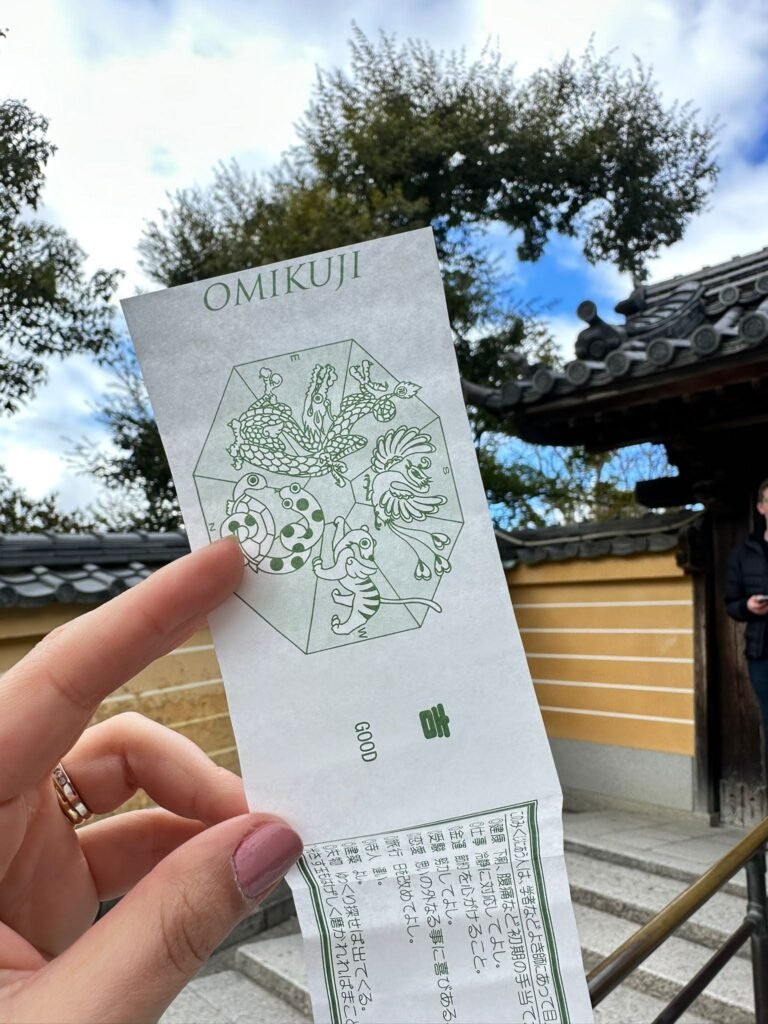 Close-up de uma mão segurando um pequeno papel de oráculo japonês (Omikuji) dobrado, com o ideograma de "Boa Sorte" visível. O fundo é suavemente desfocado, mostrando a arquitetura de madeira do Templo Kinkaku-ji (Dourado) em Kyoto.